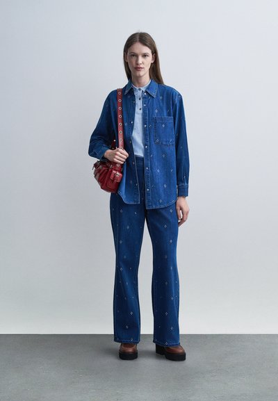 Jeune femme debout, portant une chemise et un pantalon en denim bleu assortis avec de petits motifs blancs, tenant un sac à bandoulière rouge, sur un fond de sol gris.