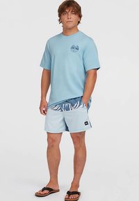 Svetlo modré tričko s krátkym rukávom, okrúhlym výstrihom a logom na prednej strane. Spárované s plastovými plavkami s vzorom a čiernymi sandálmi.
