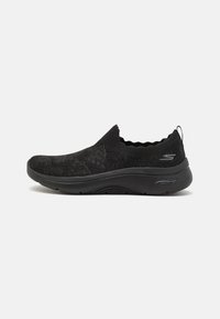 Fekete slip-on cipő, kötött felsőrésszel, textúrált mintázattal és párnázott talppal. Hullámos fodor a nyelvnél és logó dísz a oldalon.