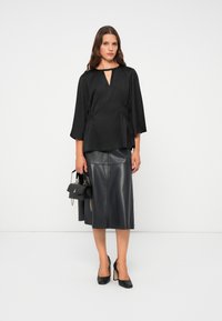 Blouse noire avec un décolleté en trou de serrure et des détails froncés, associée à une jupe midi en cuir noire et des chaussures à talons hauts noires.