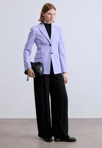 Λιλά σακάκι σε tailored γραμμή με γιακά με σχισμές, δύο μπροστινές τσέπες, μαύρη μπλούζα, φαρδύ μαύρο παντελόνι και μαύρη τσάντα με ζωνάκι. Ομαλό ύφασμα, μινιμαλιστικός σχεδιασμός.