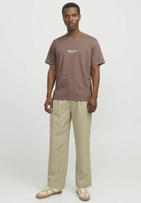 Braunes Baumwoll-T-Shirt mit Rundhalsausschnitt und kurzen Ärmeln, auf der Vorderseite mit dem weißen Text "Original Studio" bedruckt. Tragen mit beigefarbenen, gefalteten Hosen.