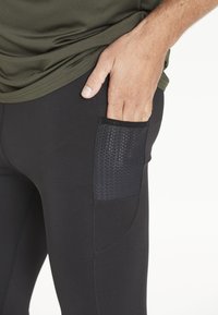 En manlig hand är inskjuten i den lilla nätfickan på sidan av svarta träningstights, iförd en olivgrön tröja.