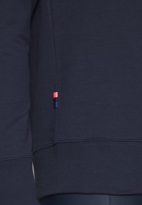 Felpa navy con orlo e polsini a coste, caratterizzata da un piccolo logo rosso, bianco e blu sul lato sinistro vicino alla cucitura.