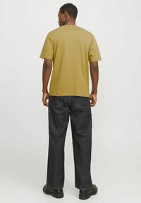 Olive green cotton t-shirt with a round neck, short sleeves, paired with black denim pants and black shoes, viewed from the back.