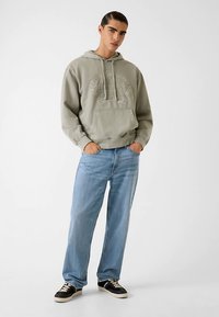 Grijze capuchontrui met reliëflogo, voorzak en trekkoorden, gecombineerd met lichtblauwe spijkerbroek en zwarte sneakers.