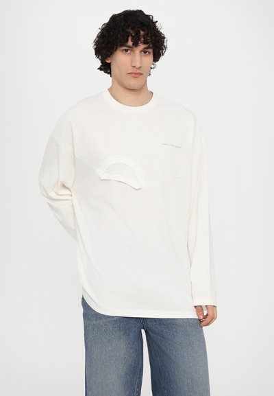 Cremfarbener Langarmshirt mit einzigartigem, geschwungenem Nahtdetail über der Brust, aus weichem Stoff, kombiniert mit blauen weiten Jeans.