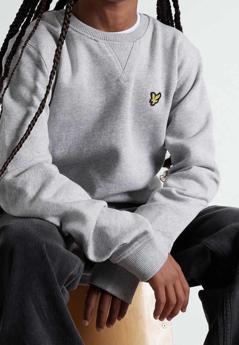 Szary bawełniany sweter z żebrowanymi mankietami i małym żółtym logo motyla na piersi. Model siedzi na drewnianym krześle, nosząc ciemne spodnie.