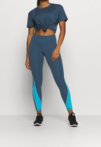 Kvinna i blå cropped träningsöverdel och matchande blå leggings med turkosa sidopaneler, stående mot en enhetlig bakgrund.