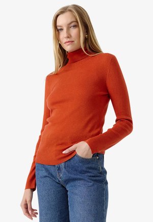 Donna con capelli biondi lunghi che indossa un maglione a collo alto arancione e jeans blu con una mano in tasca su sfondo bianco.