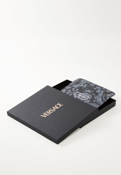 Juodas dėžutė su auksiniu "VERSACE" logotipu, iš dalies atidaryta, kad būtų matyti viduje esantis spalvingas gėlių rašto daiktas, kuris turi sidabrinius akcentus.