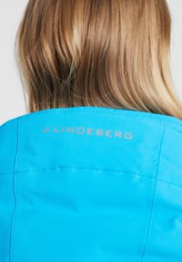 Turkos jacka med en slät textur, med subtila sömnadsdetaljer och märket "J. Lindeberg" broderat på baksidan.