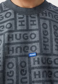 Sudadera gris con un patrón repetido de "HUGO" en tonos más oscuros, con una pequeña etiqueta azul en el dobladillo. Cuello acanalado.
