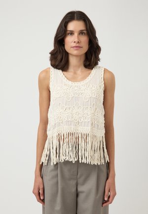 Kvinne iført hvit ermeløs heklet topp med blomster detaljer og frynser nederst, kombinert med grå høytlivsbukser, står foran en ensfarget bakgrunn.