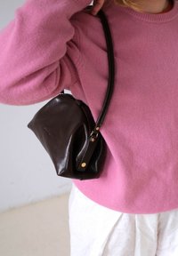 Ruda odinė rankinė su apvalia forma, reguliuojama dirželiu ir žalvarinėmis detalėmis. Dėvima kartu su rožine megztiniu ir baltais apatiniais.