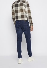 Camisa de cuadros en tonos marrón y crema, con mangas largas. Se lleva con jeans de denim oscuros y zapatillas blancas. Bolsillos traseros visibles en los jeans.