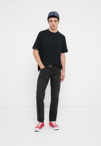 T-shirt noir, jean noir et baskets rouges. Accessoires : une casquette en denim bleu. Le modèle se tient devant un fond blanc uni.