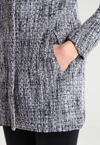 Cardigan lavorato a maglia in un motivo testurizzato bianco e nero, con tasche laterali e maniche lunghe, caratterizzato da un evidente dettaglio della trama del tessuto.