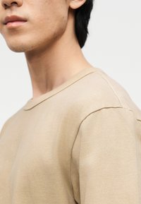 Κοντινό πλάνο ενός ατόμου που φοράει μπεζ μπλούζα με στρογγυλή λαιμόκοψη, εμφανίζοντας το κάτω μέρος του προσώπου, το λαιμό και τον ώμο σε απλό φόντο.