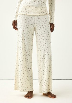 Persona che indossa pantaloni larghi color crema con piccoli motivi floreali e una maglia a maniche lunghe coordinata, in piedi a piedi nudi su un pavimento semplice.