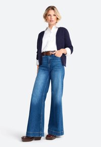 Cardigan blu navy sopra una camicia bianca, jeans in denim a vita alta e gamba ampia di colore azzurro chiaro, e scarpe con tacco quadrato marroni. Texture morbida, vestibilità casual.