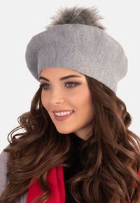 Chapeau beret en tricot gris, avec une forme arrondie, une texture douce et un pompon duveteux sur le dessus ; présente un accent de marque métallique.