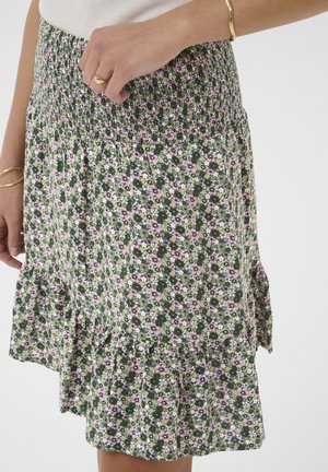 Jupe à motif floral avec des fleurs roses et blanches sur fond vert, taille élastique, portée avec une bague en or et des bracelets.