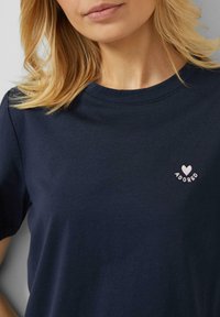 Blonde Frau, die ein dunkelblaues T-Shirt mit einem kleinen weißen Herz und dem auf der Brust gestickten Schriftzug "ADORED" trägt.