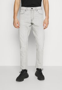 Hombre vestido con jeans slim-fit gris claro y zapatillas negras, de pie frente a un fondo blanco liso.