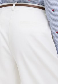 Posnetek v bližini osebe, ki nosi bele hlače z zadnjim žepom in rjavim pasom, v kombinaciji z modro srajco z modrimi črtami in vezenino cvetov na rokavu.