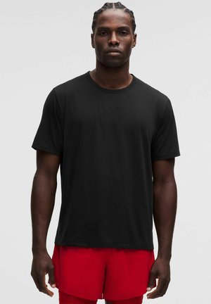 Homme aux cheveux tressés portant un T-shirt noir uni à manches courtes et un short rouge, debout devant un fond gris clair uni.