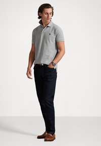 Polo gris con cuello acanalado y pequeño logo, combinado con vaqueros oscuros y mocasines marrones. Diseño simple y clásico.