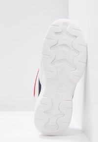 Semelle de basket blanche avec le logo FILA, motifs de grip texturés et panneaux latéraux visibles en rouge et bleu marine appuyée contre un mur blanc.