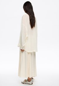 Camisa de gran tamaño en crema clara, hecha de un tejido transparente, combinada con una falda midi a juego con dobladillo desflecado. Zapatillas plateadas completan el conjunto.