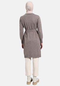 Long brown knitted cardigan with a belt at the waist, worn over cream trousers and white ankle boots. Smooth texture, subtle pattern.