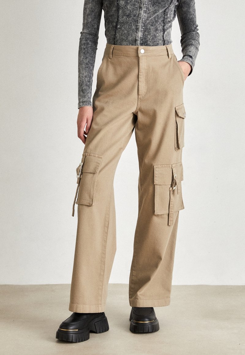 Femme portant un pantalon cargo beige avec plusieurs poches à rabat et des chaussures plateformes noires épaisses sur fond uni.
