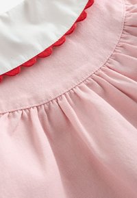 Pehme vaaleanpunainen kangas, jossa laskostettu ommel valkoisen kauluksen alla, joka on reunustettu punaisella aaltoilevalla reunuksella.