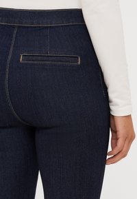 Jeans de ganga escura com um design ajustado, apresentando um único bolso traseiro e costura em contraste. O material tem uma textura suave.