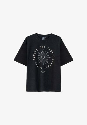 Automat sprzedający z czerwonym zewnętrzem z brandingiem Coca-Coli, otoczony różnymi wyborami napojów. Model stoi w luźnych jasnoniebieskich spodniach i czarnej koszulce.