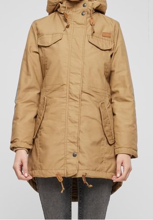 Beige hættejakke til vinteren med forlommer med klap foran, snore og lukning med trykknapper, båret med mørke jeans, klippet midt på låret.