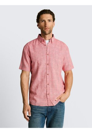Mann mit kurzen lockigen Haaren trägt ein kurzärmeliges rosa Hemd mit Knöpfen über einem weißen Unterhemd und blauen Jeans, stehend vor einem schlichten Hintergrund.