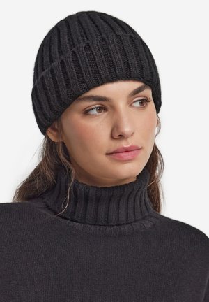 Ung kvinde iført en sort ribstrikket hue og en matchende sort ribstrikket højhalset sweater, der kigger let til højre.