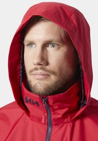 Czerwona wodoodporna kurtka z kapturem, z granatowymi akcentami i zamkiem z przodu. Zbliżenie pokazuje teksturę materiału i logo na kołnierzu.