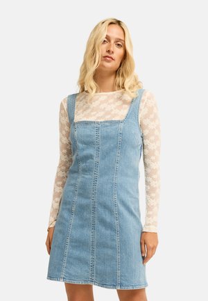 Blonde vrouw die een mouwloze blauwe spijkerjurk draagt over een lange mouw doorzichtige witte bloementop, staande tegen een effen achtergrond.