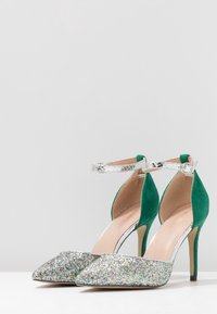 Gröna högklackade skor med silverfärgad ankelrem och glittrig silvertå, mot en enkel vit bakgrund.