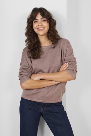 Giovane donna con capelli castani ricci, che indossa un maglione malva e jeans blu scuro, in piedi con le braccia incrociate contro una parete bianca.