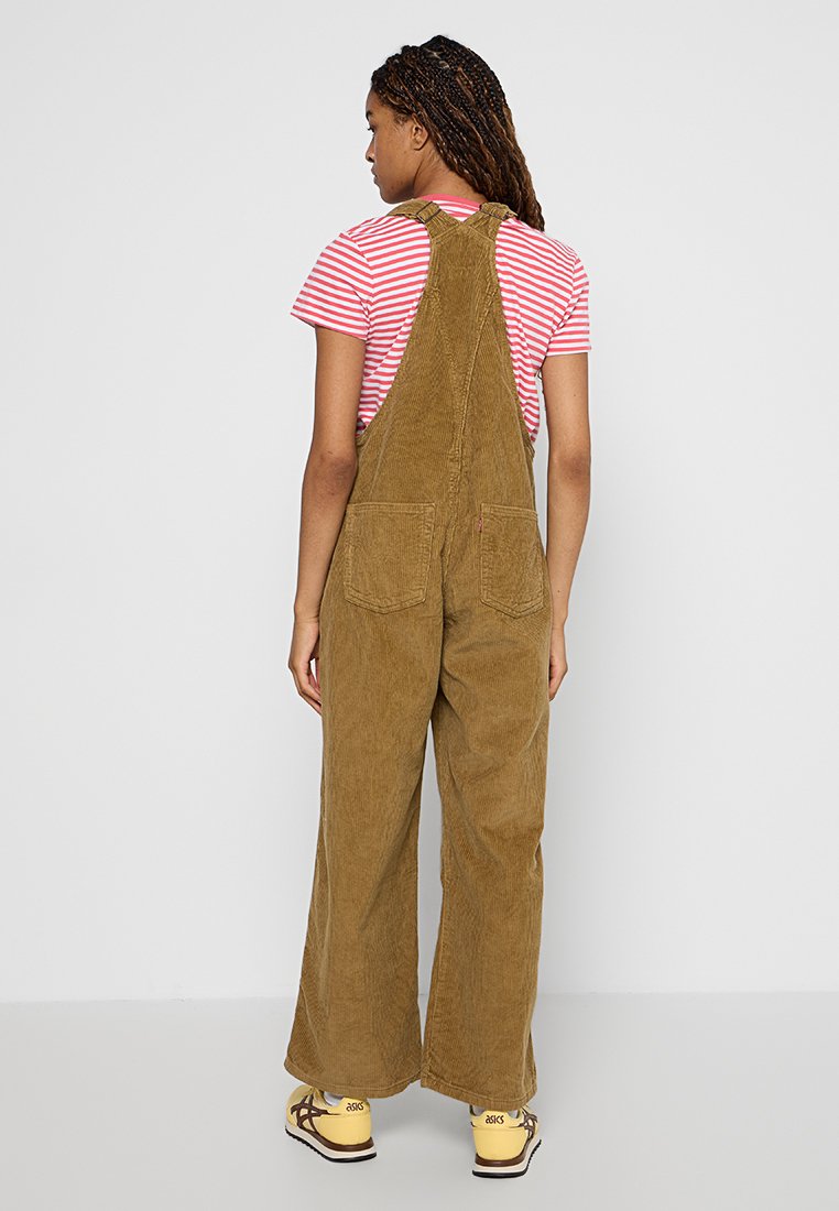 Salopette en velours côtelé marron avec des jambes larges, deux poches arrière, portée par-dessus un t-shirt rayé rouge et blanc. Le mannequin est debout avec les cheveux en tresses.