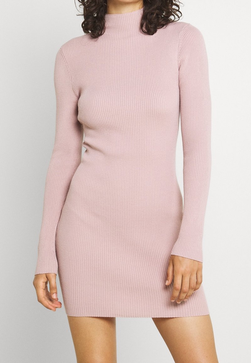 Femme portant une robe tricotée rose ajustée à manches longues côtelées, debout avec une main détendue et l'autre légèrement pliée.