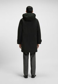 Uomo in piedi di spalle, indossa un cappotto nero con cappuccio, pantaloni grigi e scarpe nere, su uno sfondo grigio uniforme.