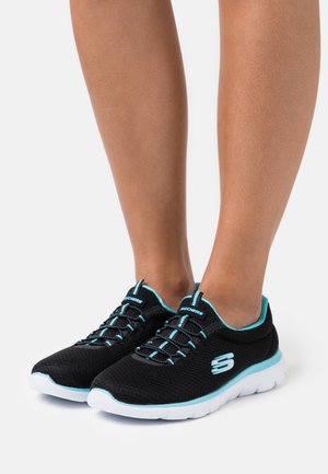 Musta Skechersin liukusovitus-tennarit sinisillä yksityiskohdilla ja valkoisilla pohjilla, jalassa paljain jaloin, kuvattu edestä ja sivulta valkoisella taustalla.